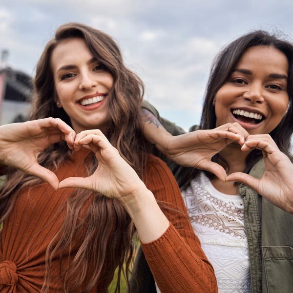 Two women smiling and making heart shapes with their hands outdoors.