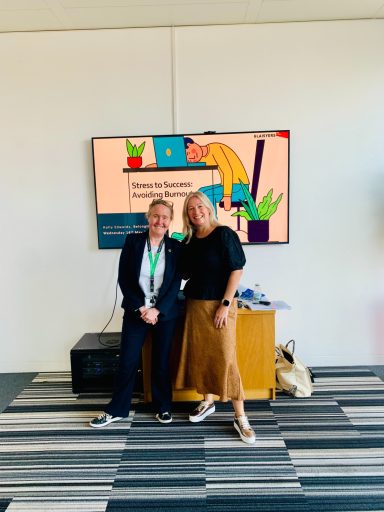 Two women standing together in an office setting, with a presentation visible behind them.