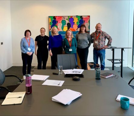 Group of six people standing in an office setting, with a colourful wall art behind them.