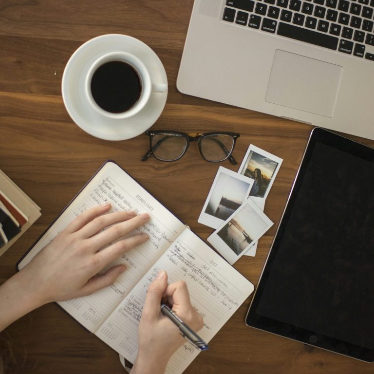 A person's hands writing in a notebook, with coffee, glasses, and photos on a wooden table.