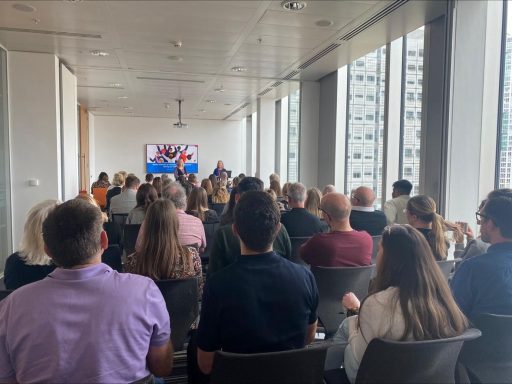 A crowd of people seated in a modern meeting room, facing a presentation screen.
