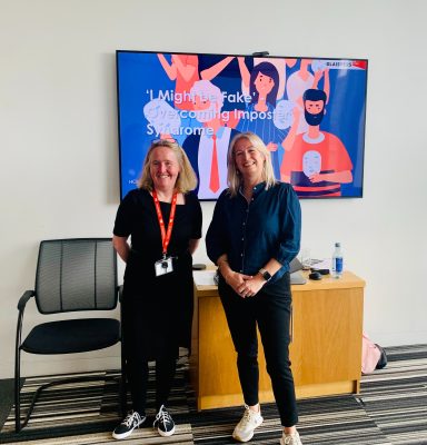 Two women stand smiling in front of a digital display featuring illustrations of diverse people.